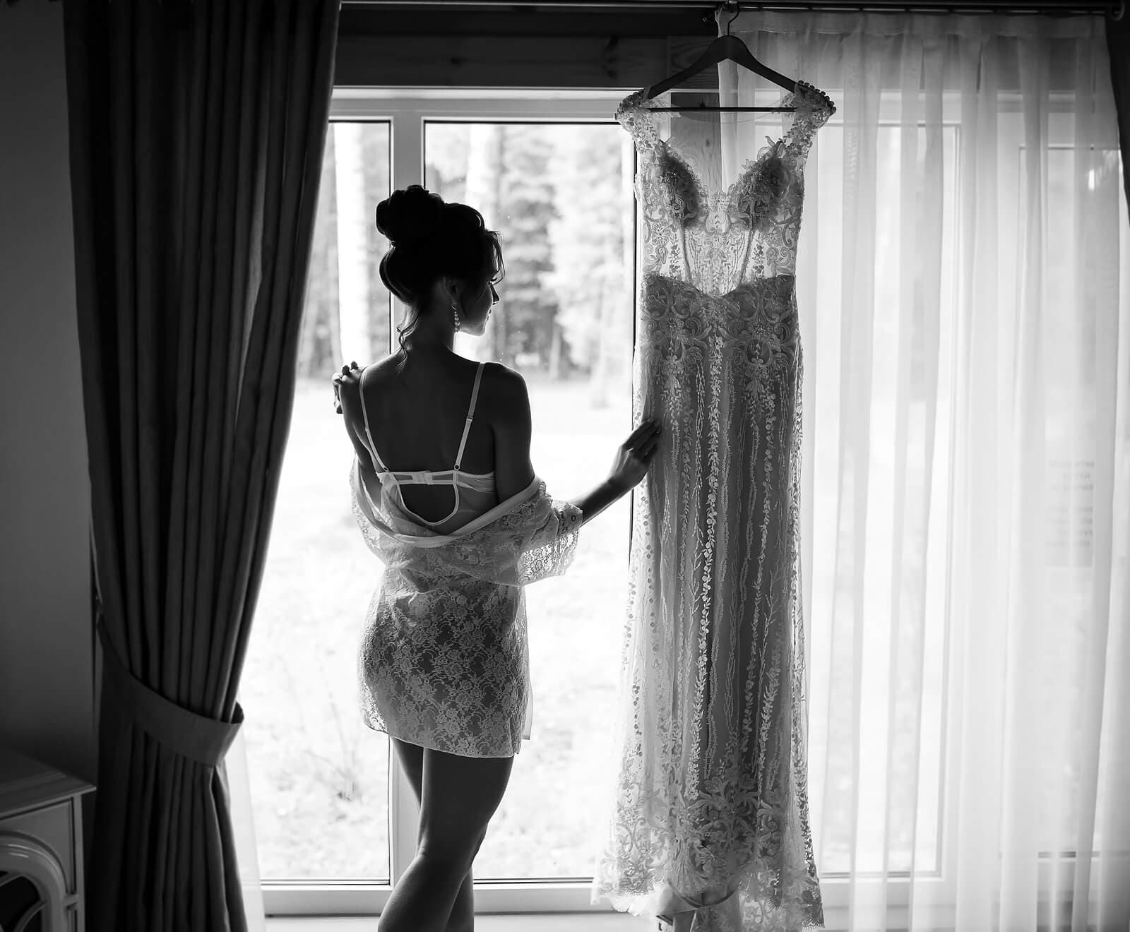 Bride in lace gown beside a wedding dress hanging in the window